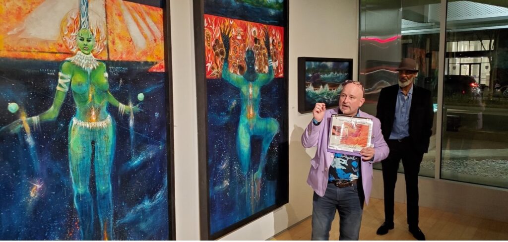 Jason Lye stands in front of two paintings of African deities, featuring orange and yellow shades.  Jason is explaining that clay-based orange and yellow pigments and an ancient paint factory was discovered in Africa, dated 150,000 years ago.