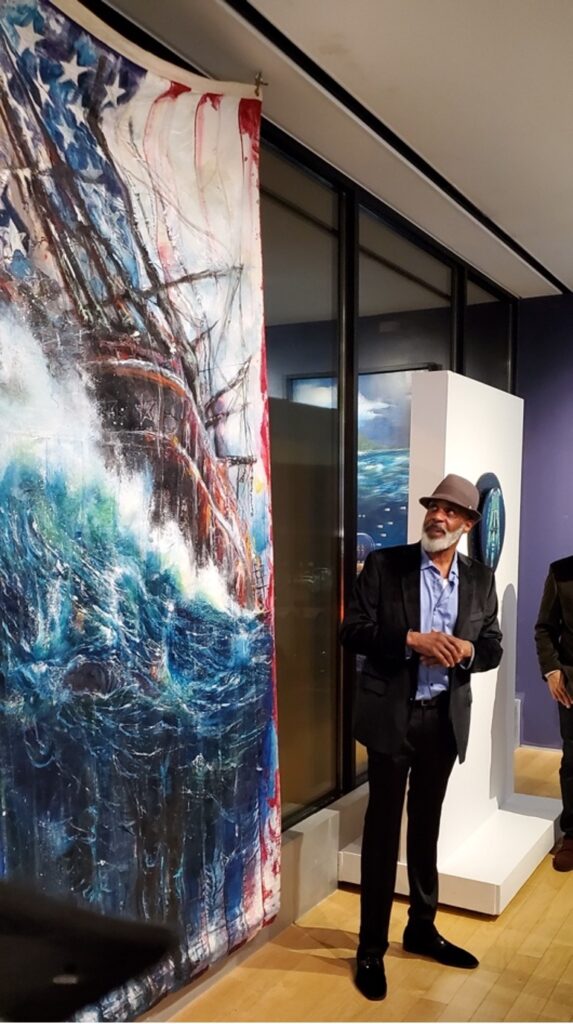 Artist Andre Henderson in front of a rear-view image of the Clotilda (last slave ship) on a rough sea.  The image is painted on a stitched US flag.
